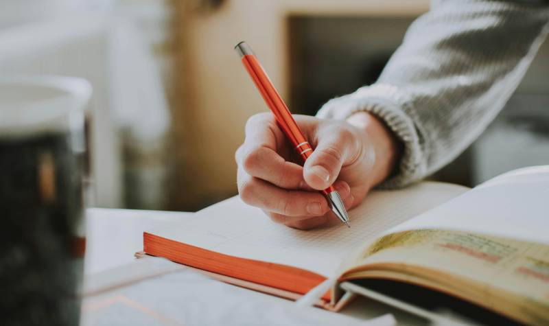 Person writing with an orange pen on a notebook next to a glass of water.