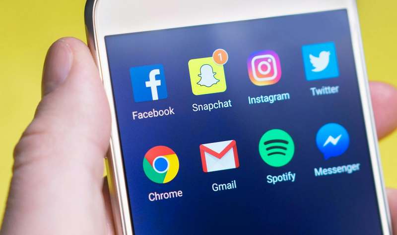 A hand holds a smartphone displaying popular social media and communication apps like Facebook, Snapchat, Instagram, Twitter, Chrome, Gmail, Spotify, and Messenger.