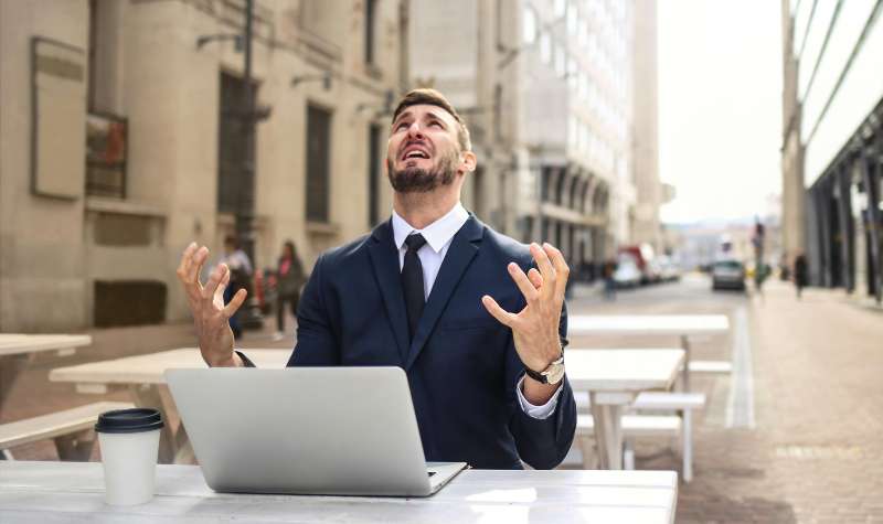 Man in suit at outdoor cafe, hands raised, looking up with laptop and coffee cup nearby.