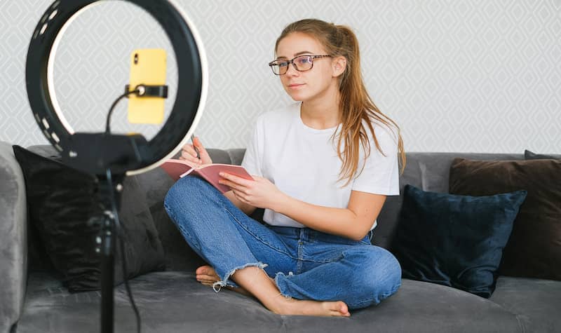 Woman recording video with smartphone on ring light while sitting on couch.