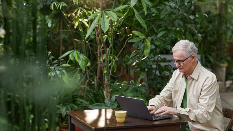 Man working on laptop at outdoor table with greenery around.