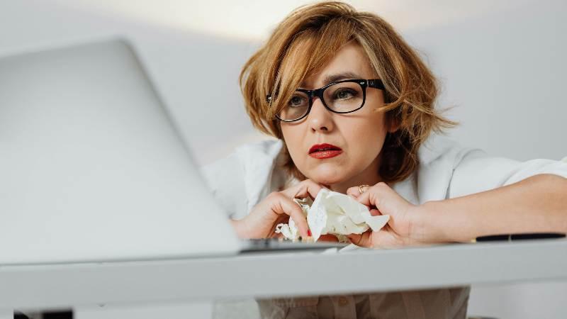 Woman at desk with laptop, holding crumpled paper, looking thoughtful.