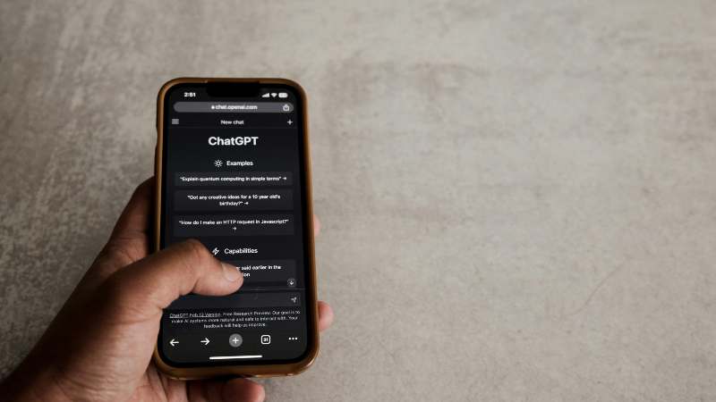 A hand holding an iPhone displaying ChatGPT app interface with examples and capabilities.