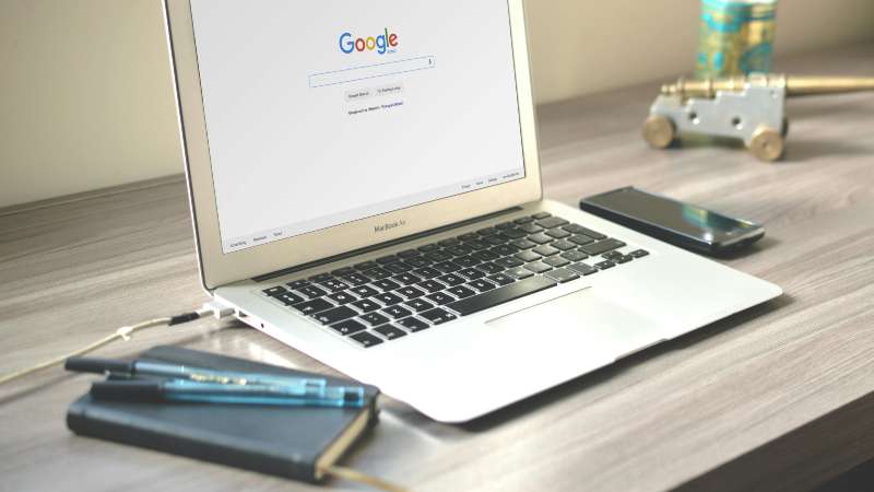 Laptop with Google homepage open on desk, alongside smartphone and pens.