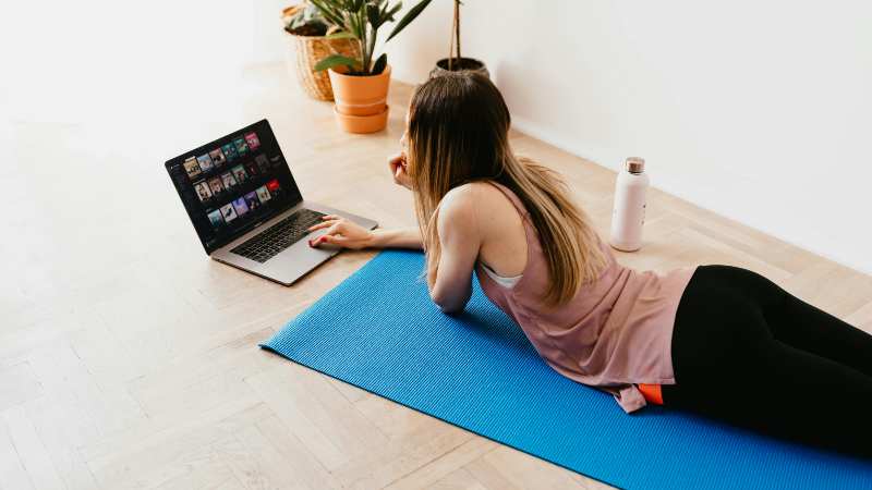 Woman lying on yoga mat using laptop with plants nearby.