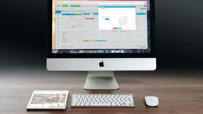 Apple iMac with keyboard and mouse, tablet on desk, displaying a scheduling software interface.