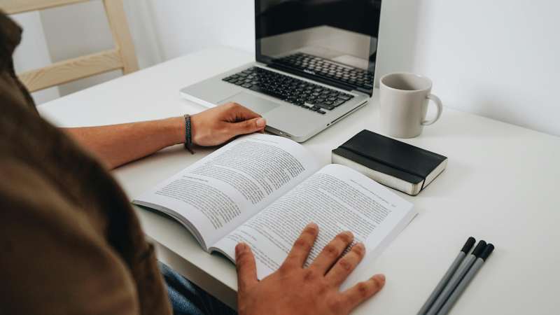 Person studying at a desk with an open book, laptop, notebook, pen, and coffee cup.