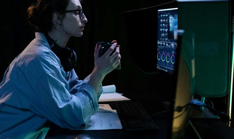Woman working intently at computer with headphones, holding cup, surrounded by technical equipment.
