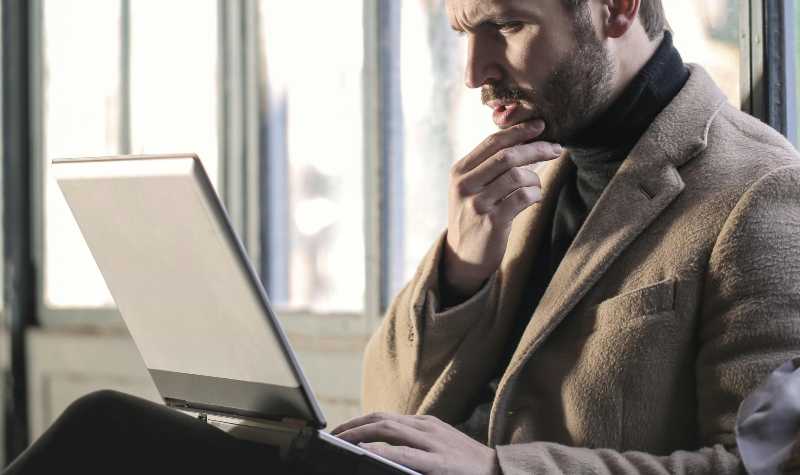 Man in brown coat using laptop by window.
