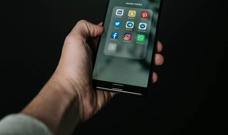 A hand holds a smartphone displaying popular social media apps like LinkedIn, Twitter, Facebook, Instagram, WhatsApp, Pinterest, Snapchat, and Messenger.