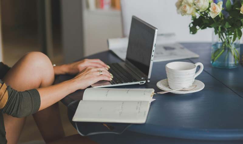 Woman typing on laptop with notebook and coffee nearby.