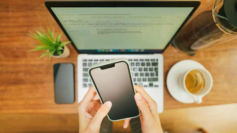 Hands holding a smartphone with laptop and coffee cup nearby.