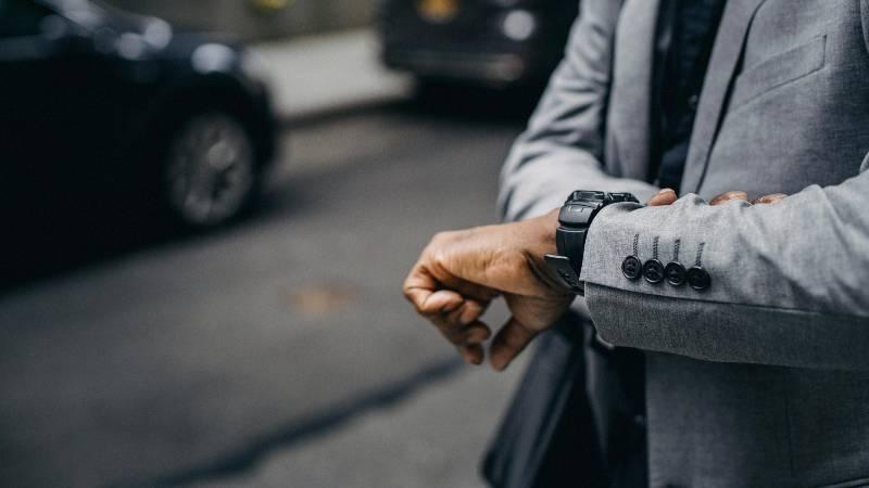 Man in gray suit checking his watch while standing on a street with cars passing by.