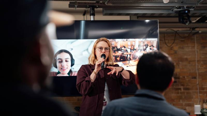 A presenter gestures while speaking to an audience, with a large screen showcasing a group discussion in the background.