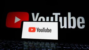 A laptop screen displays the YouTube logo, with a larger, blurred version projected behind it. The scene is dark, highlighting the bright logo in red and white.