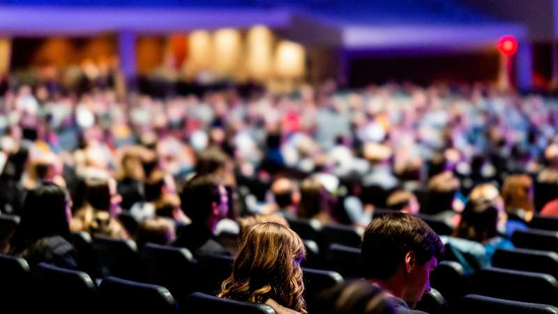 A large audience sits in a dimly lit auditorium. Seats are filled, and the crowd's attention is focused forward, creating an anticipatory atmosphere.