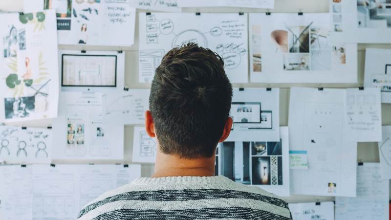 A man in a striped sweater intently examines a wall covered with sketches, graphs, and photos, suggesting brainstorming and planning in a creative environment.