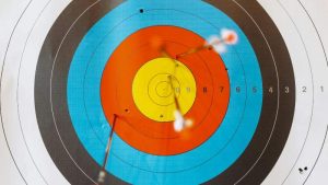 A colorful archery target with concentric circles in yellow, red, blue, and black has several arrows hitting near the center, conveying precision and focus.