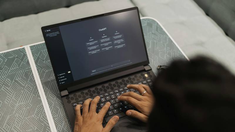 A person types on a laptop displaying the ChatGPT interface. The setting is casual, with a patterned table and sofa, conveying a focused, tech-savvy atmosphere.