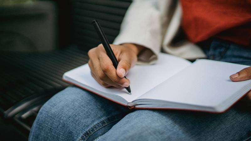 A person in jeans writes in an open notebook with a black pen, seated on a bench. The scene conveys focus and introspection.