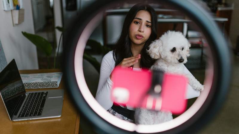 A woman holds a small white dog, speaking into a smartphone mounted on a ring light. A laptop is open on a table beside her, conveying a content creation scene.