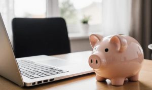 A piggy bank sits beside a laptop on a wooden desk. Natural light filters through a window, creating a peaceful workspace atmosphere.