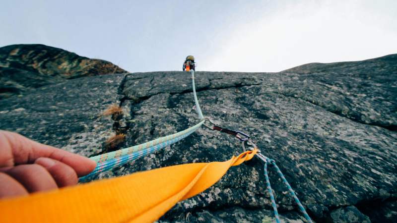 Climber ascends a steep rock face, secured by ropes and a clip, while a hand grips a bright yellow strap in the foreground, conveying adventure.