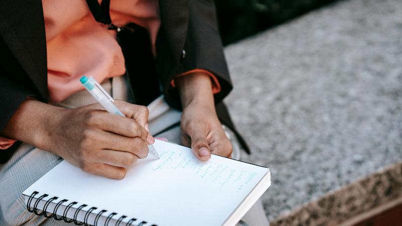 A person sits outdoors on a stone bench, writing in a notebook with a green pen. They wear a brown blazer and coral shirt, conveying focus and contemplation.
