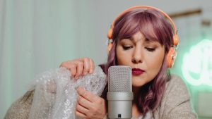 Woman with purple hair wearing orange headphones, holding bubble wrap near a microphone. She appears focused, suggesting a calm ASMR recording session.