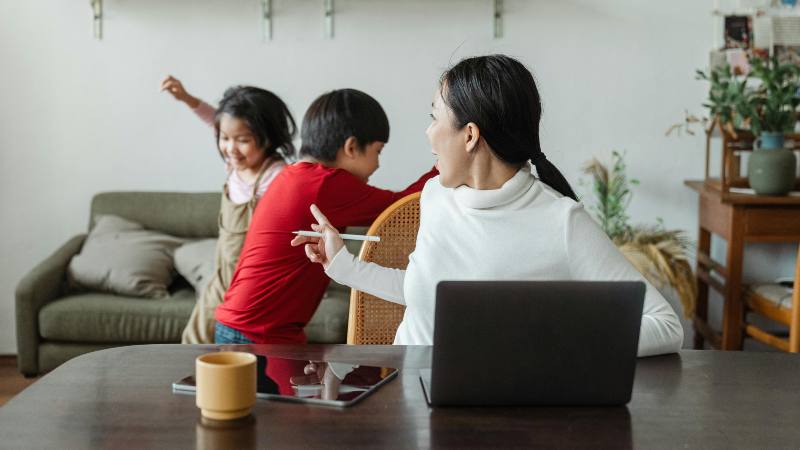 A woman working at a table with a laptop and tablet turns to watch two children playing energetically in the background, creating a lively home atmosphere.