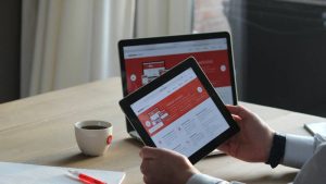 A person holds a tablet showing a website in front of a laptop displaying the same site. A cup of coffee and a notepad with a pen are on the table.