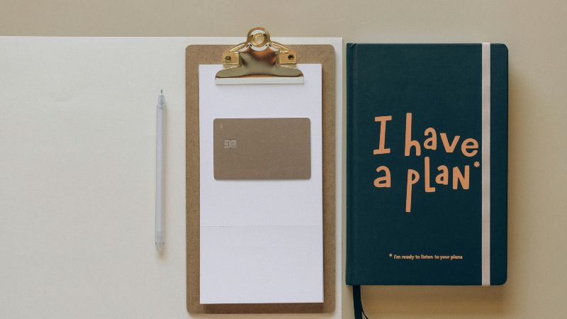 A clipboard with a card, a pen, and a dark green notebook with "I have a plan" in bold writing on it. The layout feels organized and motivational.