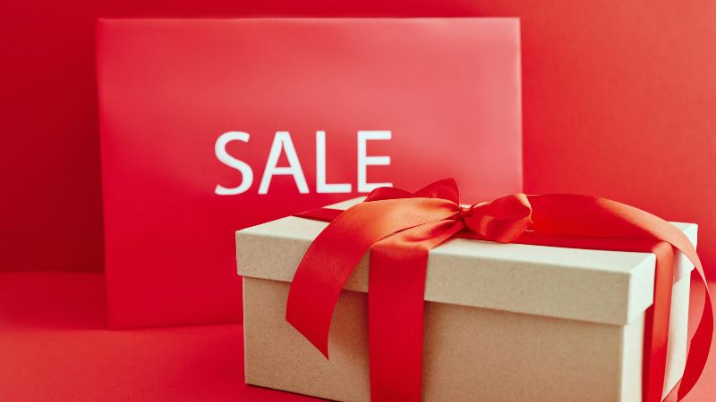 Gift box with a red ribbon in front of a red "SALE" sign. The vibrant colors convey a festive, enticing atmosphere, suggesting discounts or promotions.
