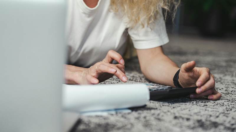 A person lies on a carpeted floor, using a calculator and looking at papers. They appear focused, with a laptop partially visible in the foreground.