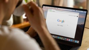 A person sips from a mug while viewing the Google homepage on a laptop. The screen displays search options, conveying a focused and work-like atmosphere.