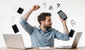 A man sits between two laptops, holding a smartphone, surrounded by floating devices and question marks, conveying confusion or multitasking in a tech setting.