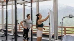 A woman writes on a whiteboard in a gym with mountain views while a man in athletic wear observes. The setting is bright and focused.
