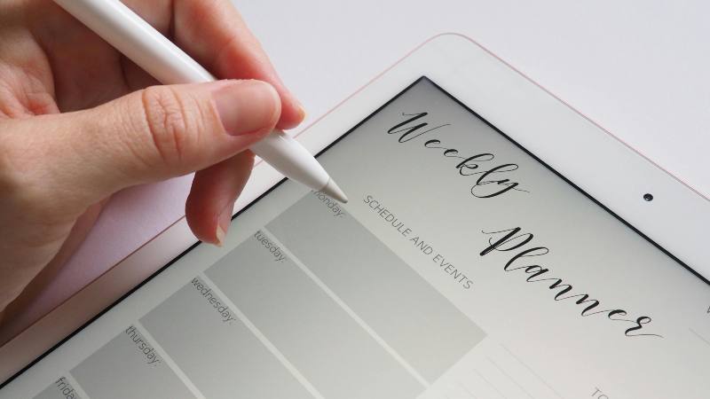 A hand holding a stylus hovers over a digital tablet displaying a "Weekly Planner" with a minimalist layout. The scene conveys organization and planning.