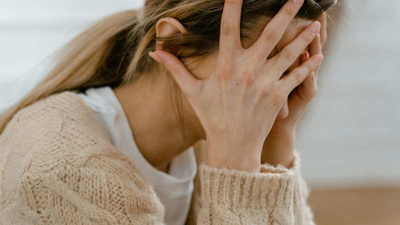 A woman with light brown hair in a ponytail is sitting, wearing a beige knitted sweater and white shirt. She covers her face with her hands, expressing stress.
