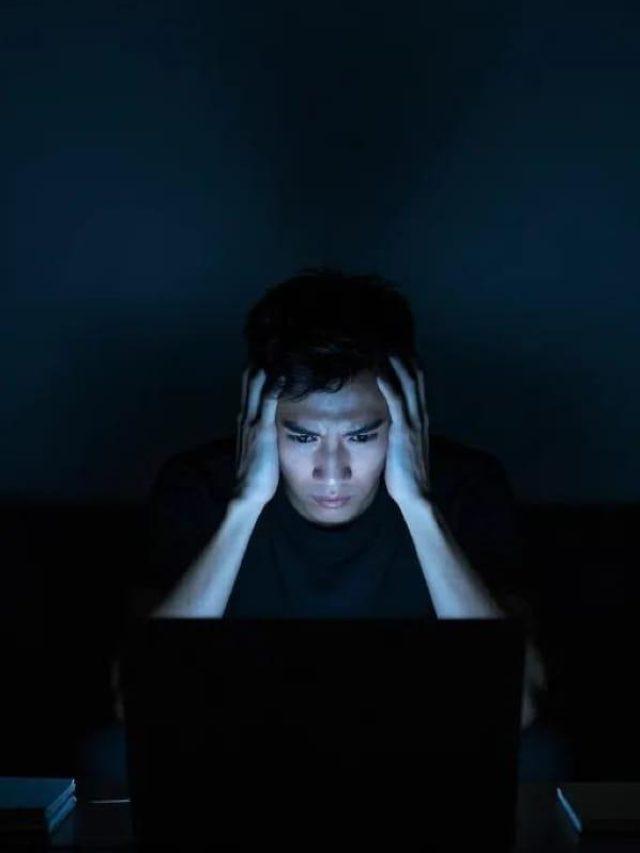 A person sitting in front of a laptop in a dark room, with their hands on their head, looking stressed and focused on the screen's glow.