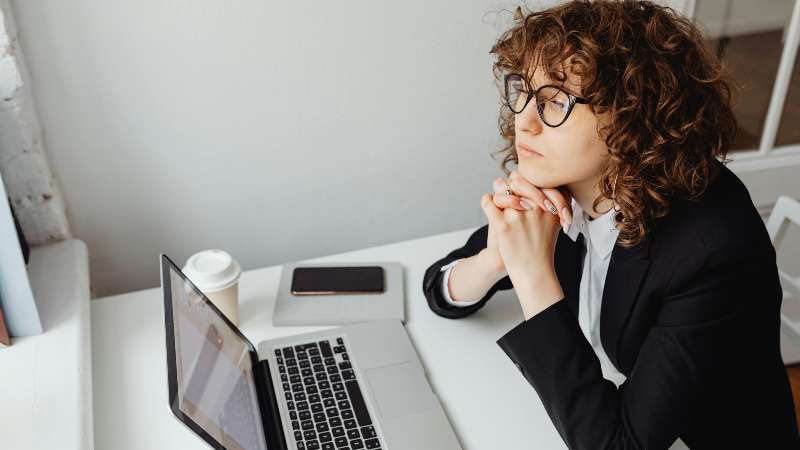 A woman with curly hair and glasses sits thoughtfully at a desk with a laptop, phone, and coffee. She wears a suit, conveying a professional tone.