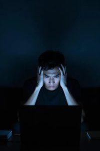 A person sitting in front of a laptop in a dark room, with their hands on their head, looking stressed and focused on the screen's glow.