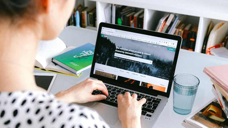 A person works on a laptop displaying a stock photo website. The desk also holds books, a notebook, and a glass of water, conveying a focused work environment.
