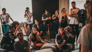 A group of people sit and stand attentively in a dimly-lit room. Some hold cameras, suggesting a workshop. The atmosphere is focused and engaged.