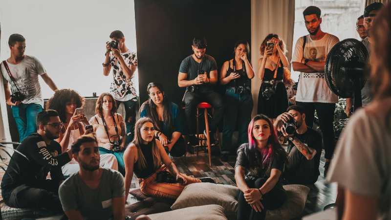 A group of people sit and stand attentively in a dimly-lit room. Some hold cameras, suggesting a workshop. The atmosphere is focused and engaged.