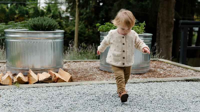 Toddler walking on gravel near planters with woodpile.