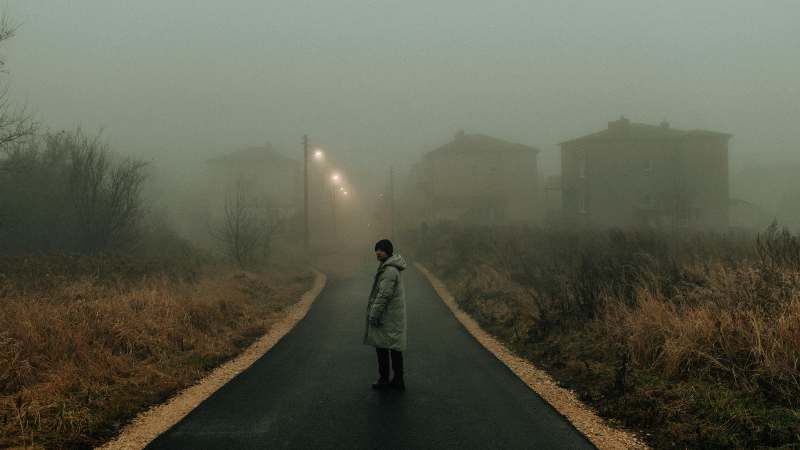A solitary figure stands on a foggy path, with houses faintly visible in the background.