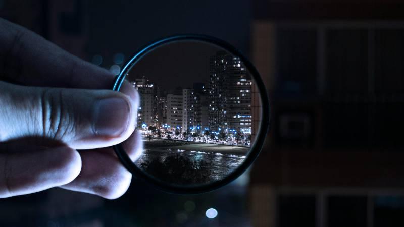 Hand holding a circular lens shows a sharp cityscape with lit buildings at night, contrasting with the blurred dark surroundings, creating a focused and mysterious tone.