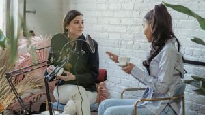 Two women are seated in a cozy room with a white brick wall, chatting over tea. A microphone on a stand suggests a podcast recording.