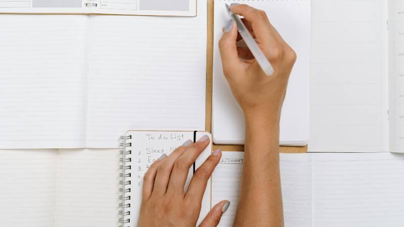 Hands write in a notebook on a desk with open planners. One list reads "To do list," followed by tasks. The scene is organized and focused.